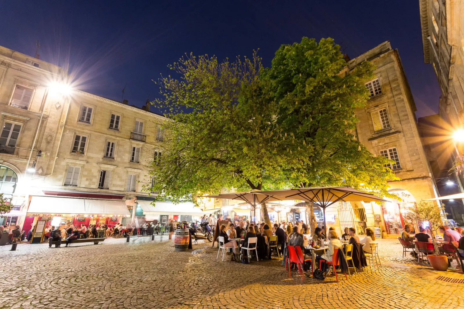 Saint-Pierre church square in Bordeaux at night, featuring illuminated restaurant terraces, historic buildings, a large tree in the center, and crowds of diners and visitors