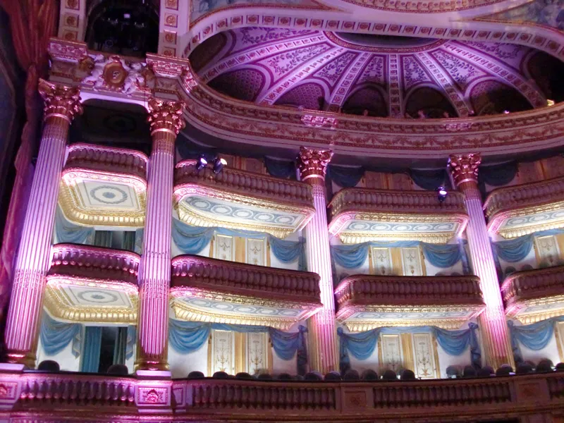 Interior view of the Grand Théâtre of Bordeaux lit in pink, displaying ornate boxes with Corinthian columns, blue and gold draperies, and the architectural dome of the ceiling.