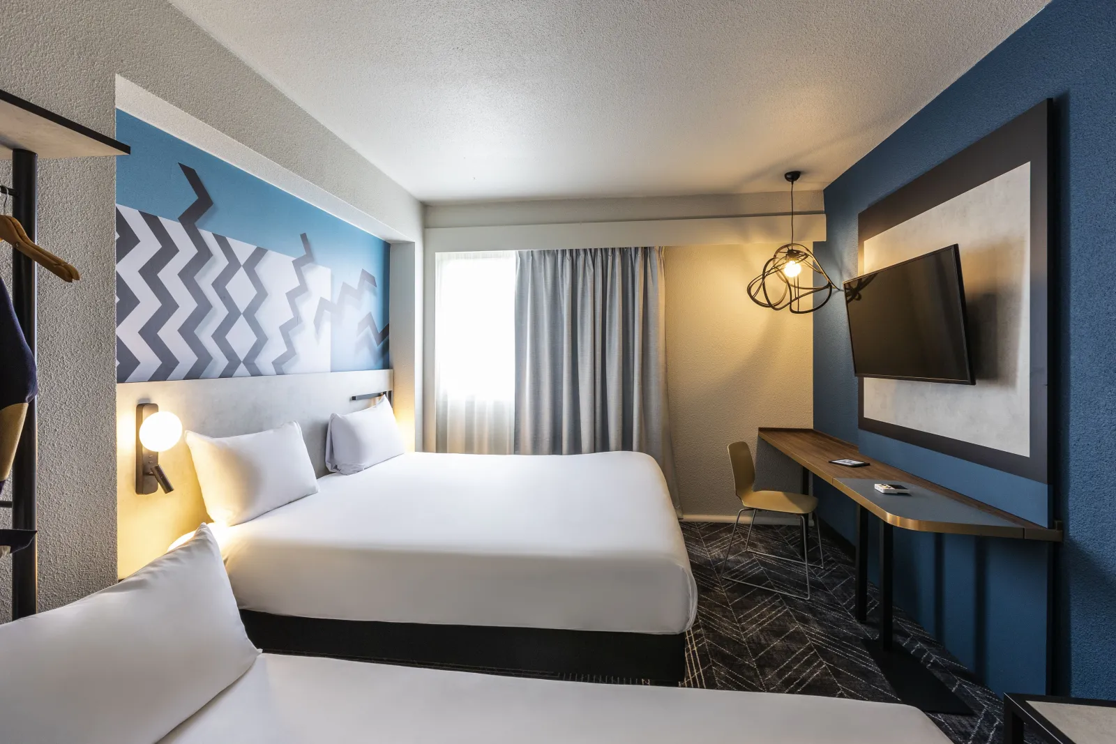 Modern double room at Ibis Styles Bordeaux Meriadeck hotel featuring two white beds, blue and white walls, desk with TV screen, and contemporary geometric decor