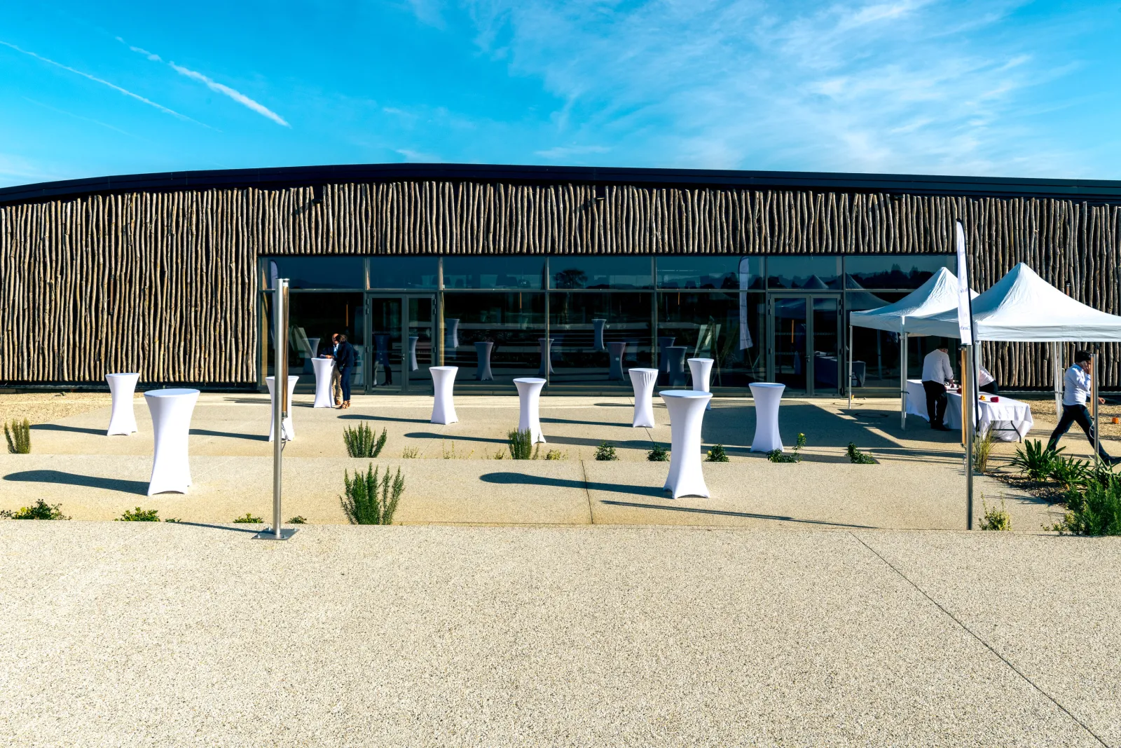 Outdoor event at the modern Château d'Arche building in Bordeaux, featuring white high-top tables, white tent structures, and attendees against a clear blue sky.