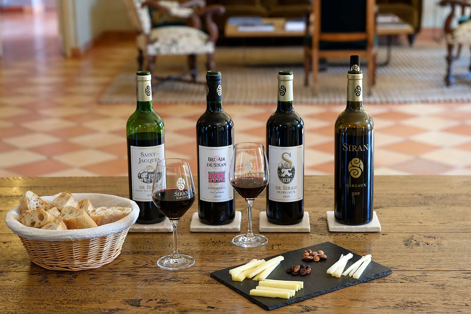 Wine tasting of Château Siran wines from Bordeaux featuring four bottles of Margaux, two filled glasses, a bread basket, and a cheese and nuts board on a wooden table.