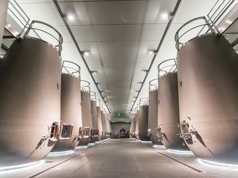 Modern interior of a wine cellar featuring two symmetrical rows of large stainless steel fermentation tanks with LED lighting, polished floor, and metal ceiling at Château Prieuré-Lichine in Bordeaux.