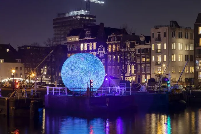 Giant illuminated blue sphere art installation on Bordeaux waterfront at night, surrounded by historic buildings lit in purple, reflected in the river water.