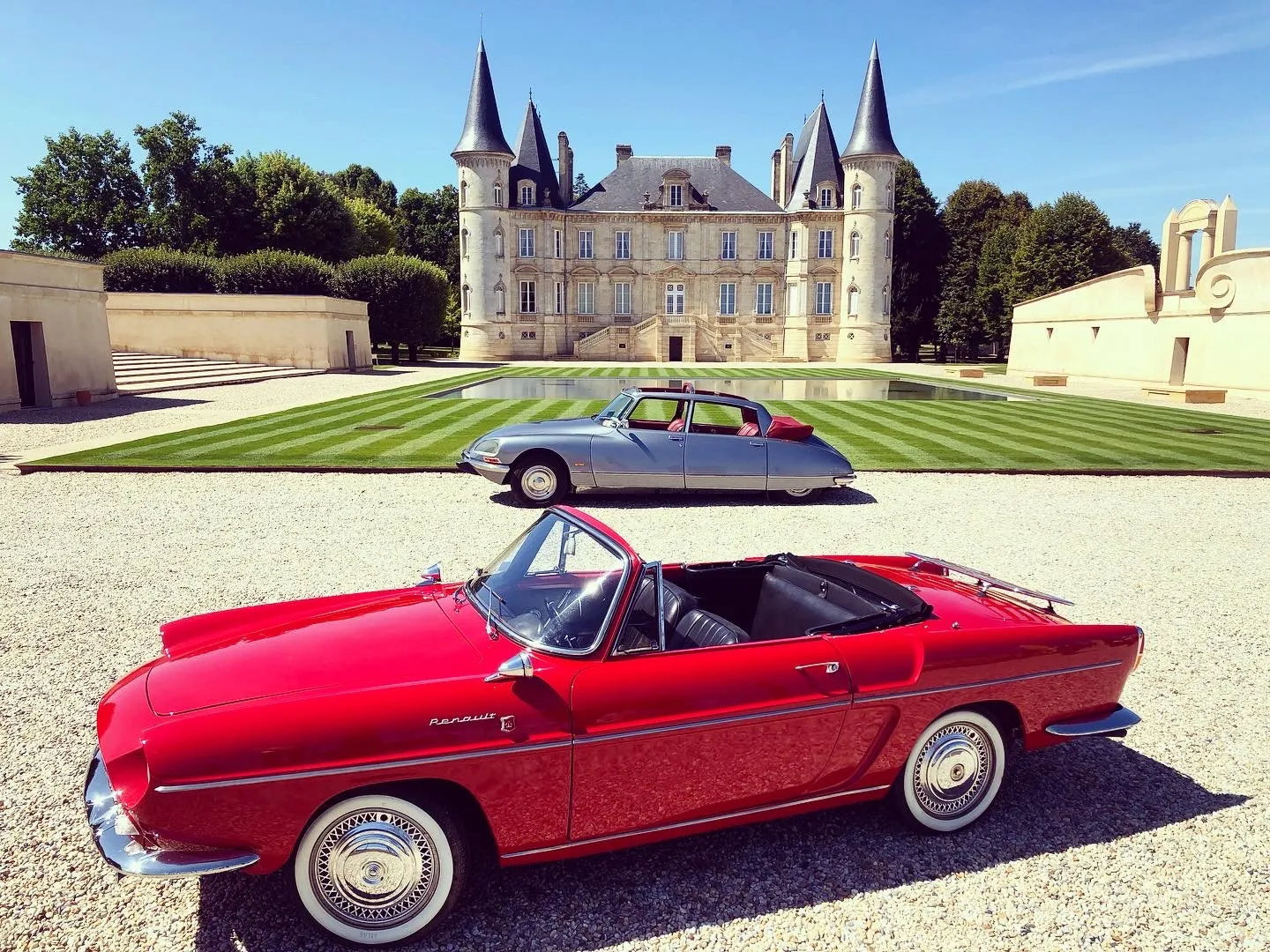 Two classic cars displayed in front of a French château with turrets: a grey Citroën DS in the background and a red convertible from the 1950s in the foreground, parked on a gravel driveway with striped lawn.