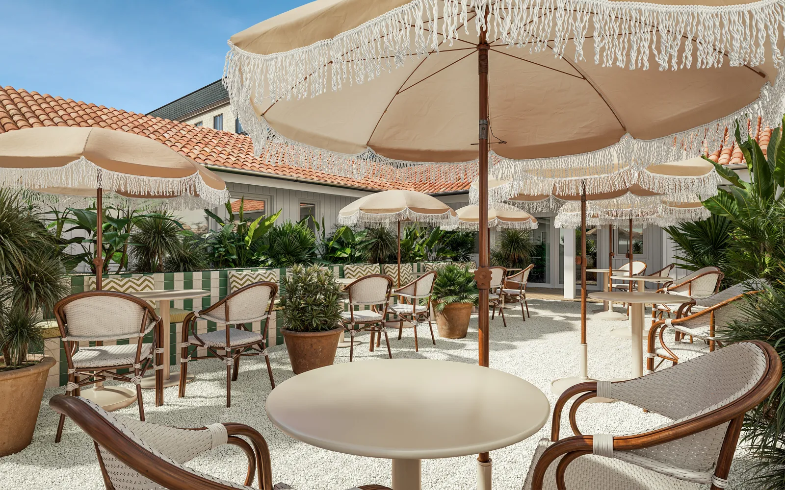 Elegant terrace at Clos Huguerie in Bordeaux featuring beige fringed umbrellas, rattan chairs, round tables, potted plants and a building with red tile roof under clear blue sky