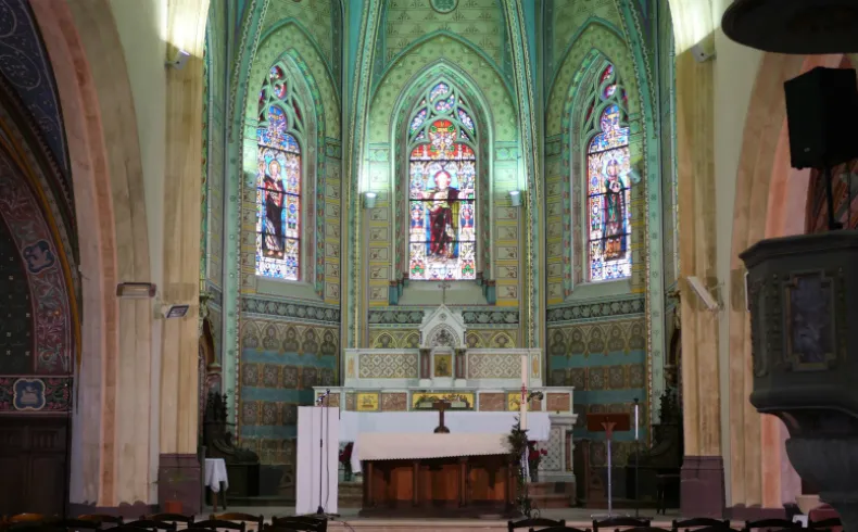 Interior of a church featuring three colorful stained glass windows above a white altar, with green ornamental decoration, in Lormont near Bordeaux.