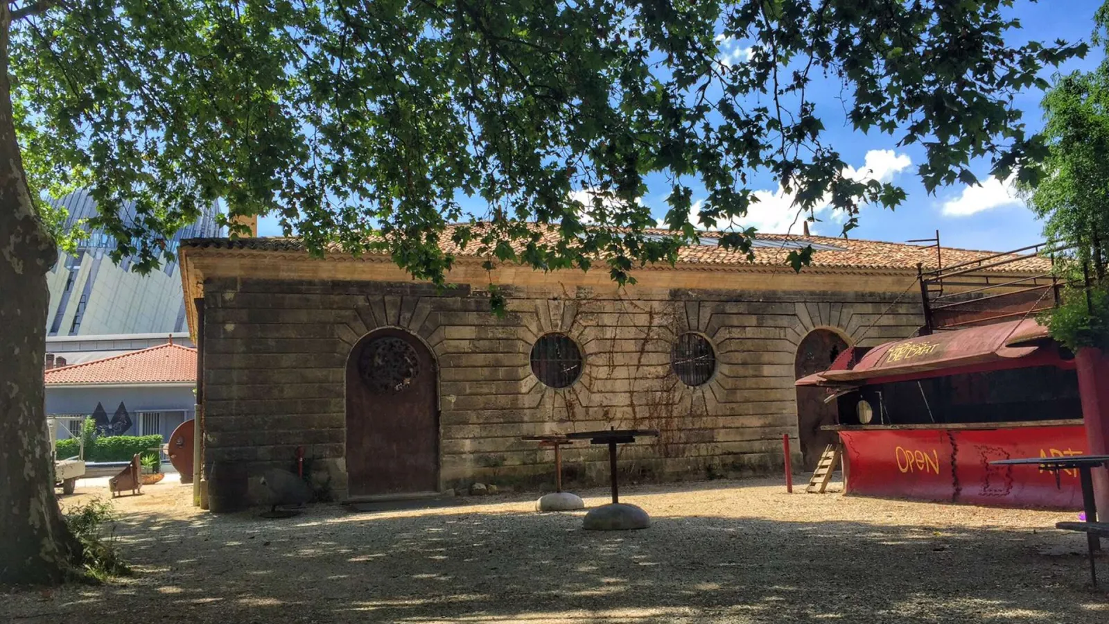 Stone building with rustic facades featuring arched doors and circular windows, next to a red metal structure marked 'OPEN', in the courtyard of Les Vivres de l'Art in Bordeaux.