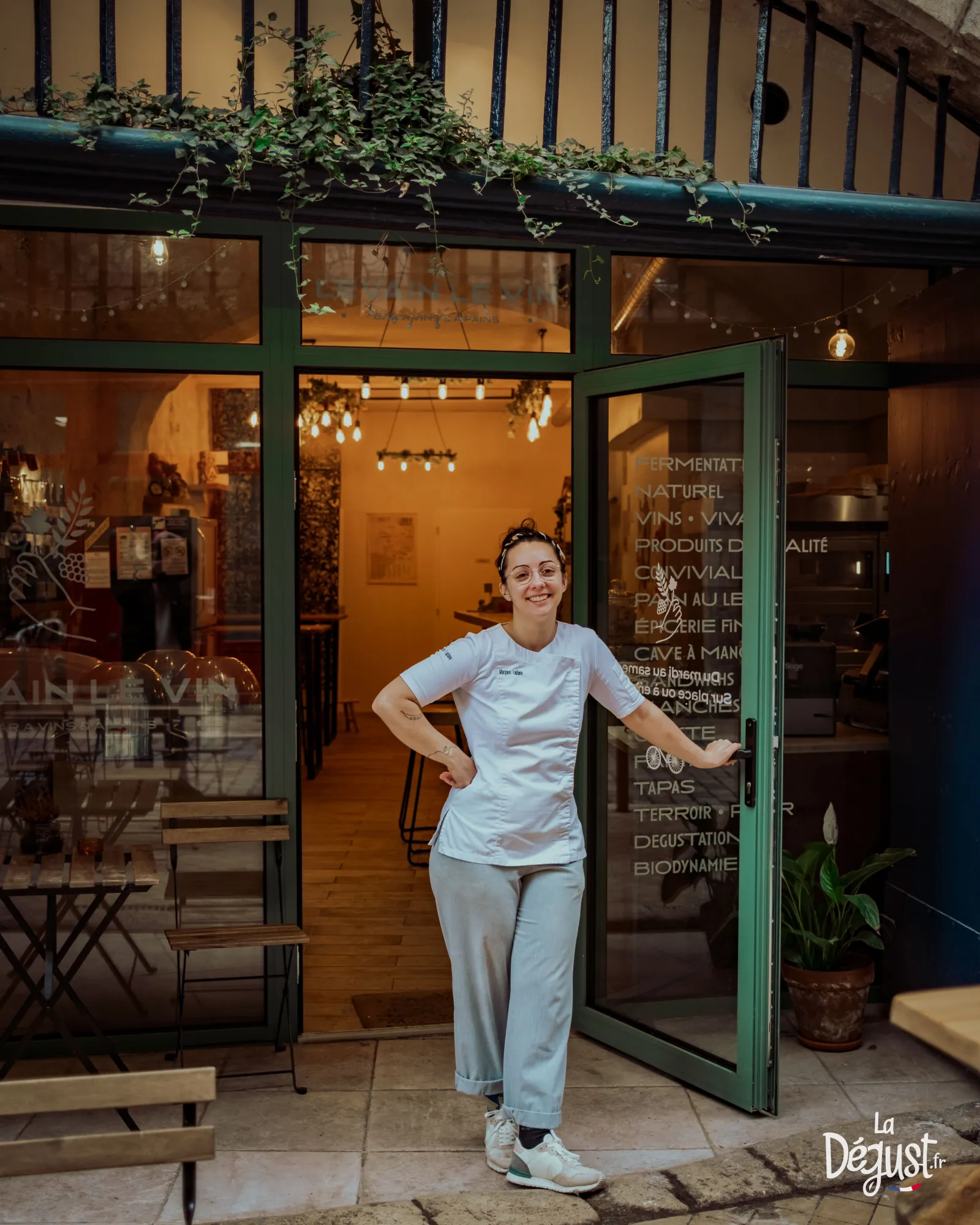 A smiling woman stands at the entrance of Levain Le Vin shop in Bordeaux, featuring green storefront and glowing windows.