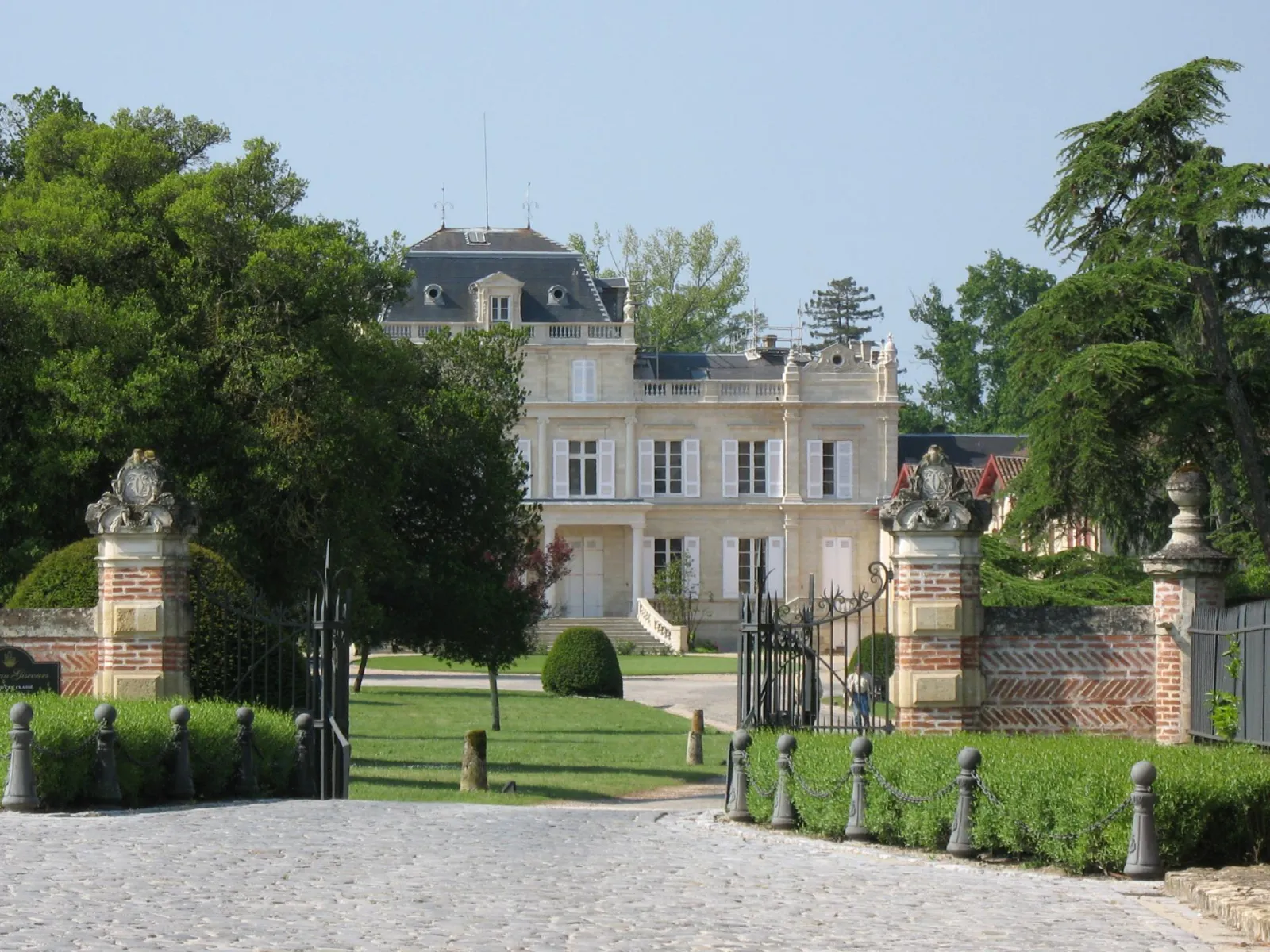 Front view of Château Giscours in Bordeaux, a neoclassical mansion with light-colored facades and mansard roofs, framed by stone gateways with sculptured busts and tree-lined pathways.