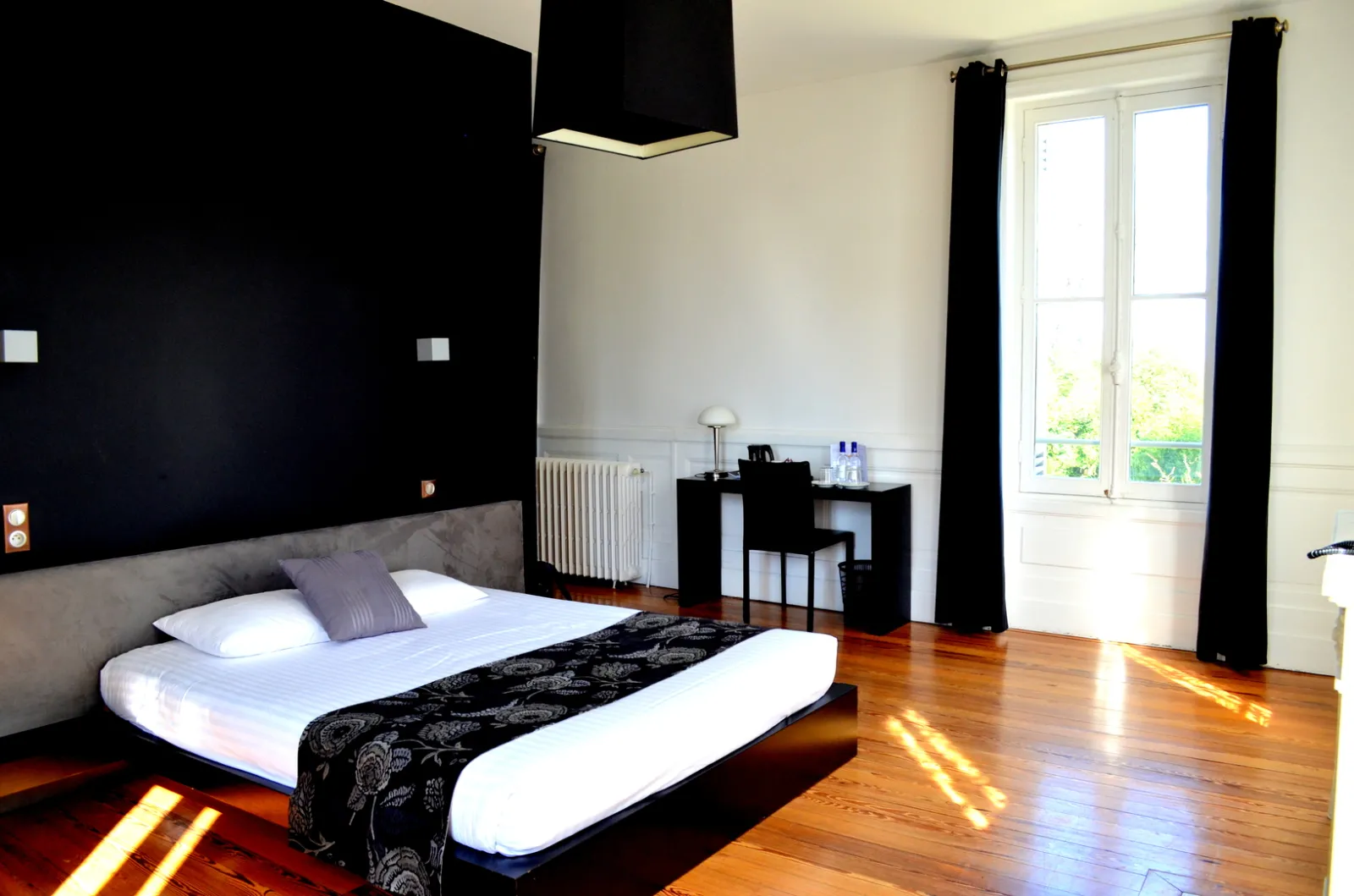 Modern and minimalist bedroom at Château La Chèze in Bordeaux, featuring a king-size bed with white linens, black and beige walls, wooden flooring, French doors opening to a garden, and black minimalist furniture.