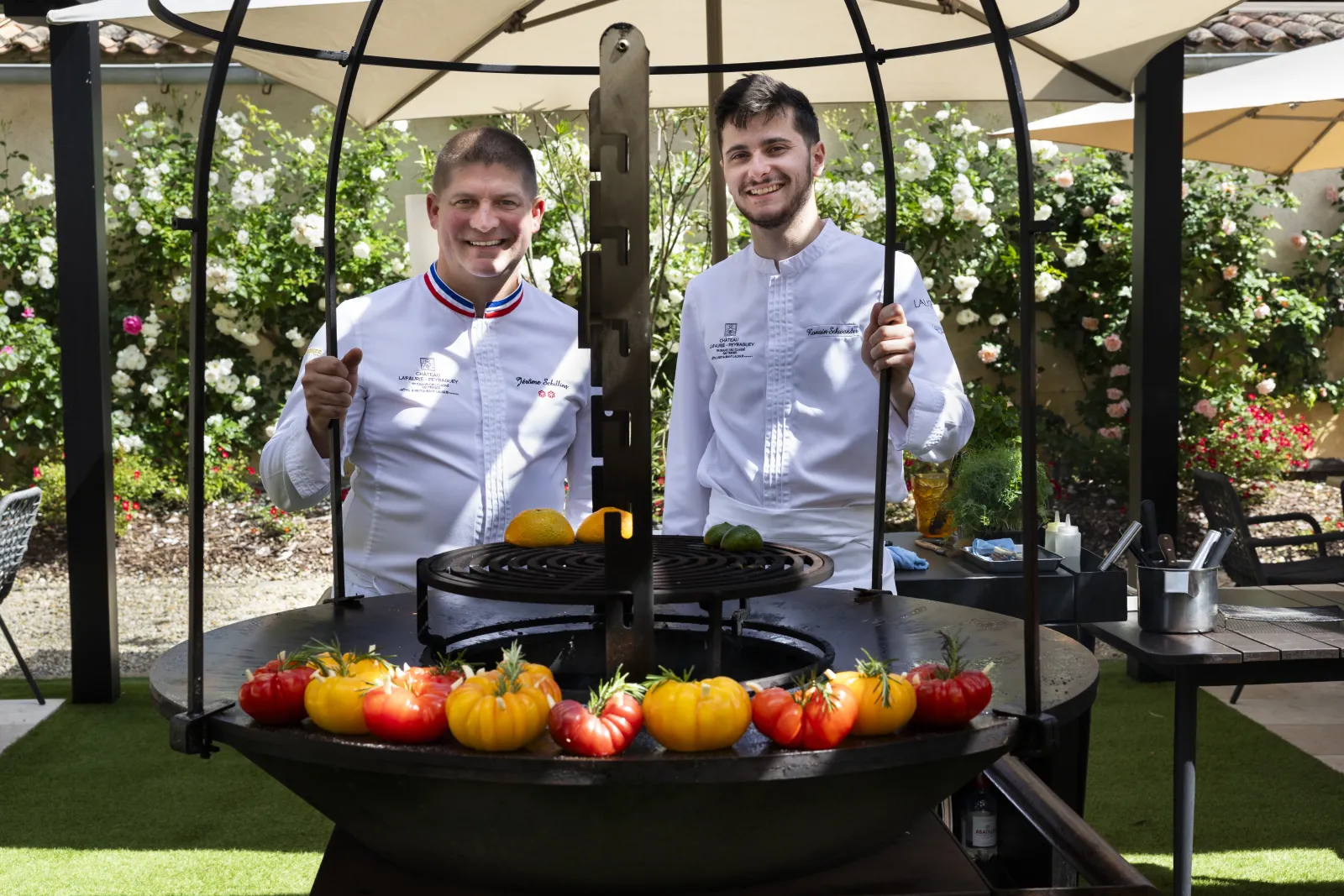 Two chefs in white uniforms pose in front of a circular brazier filled with colorful tomatoes in a flowering garden with pergola in Bordeaux.