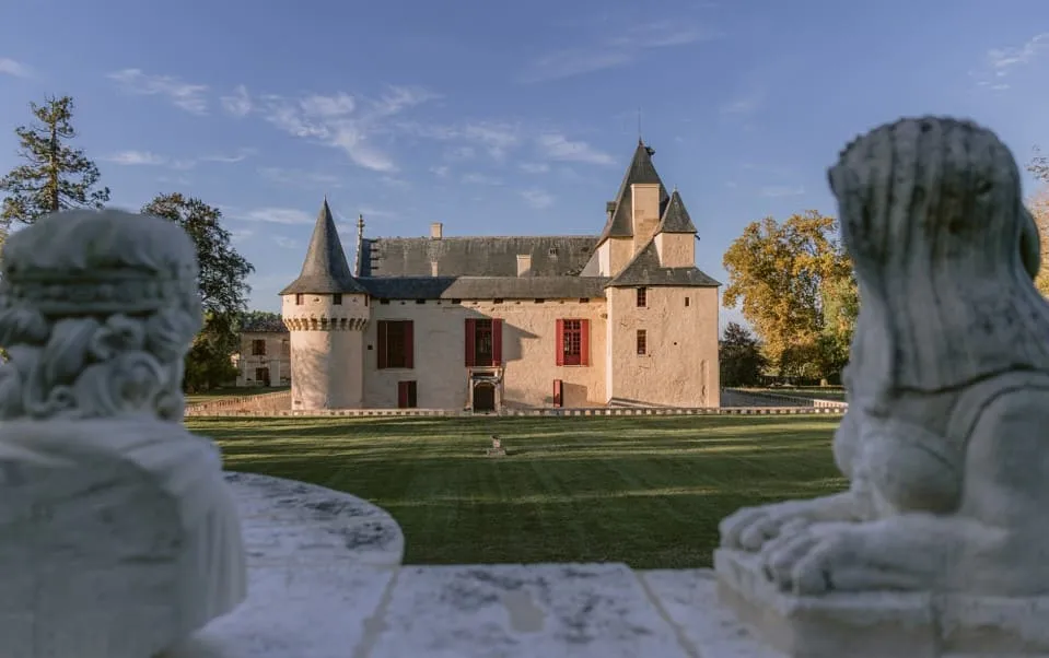 Château Olivier in Bordeaux, medieval fortress with round towers, beige facade and slate roof, framed by stone statues and surrounded by green lawn.