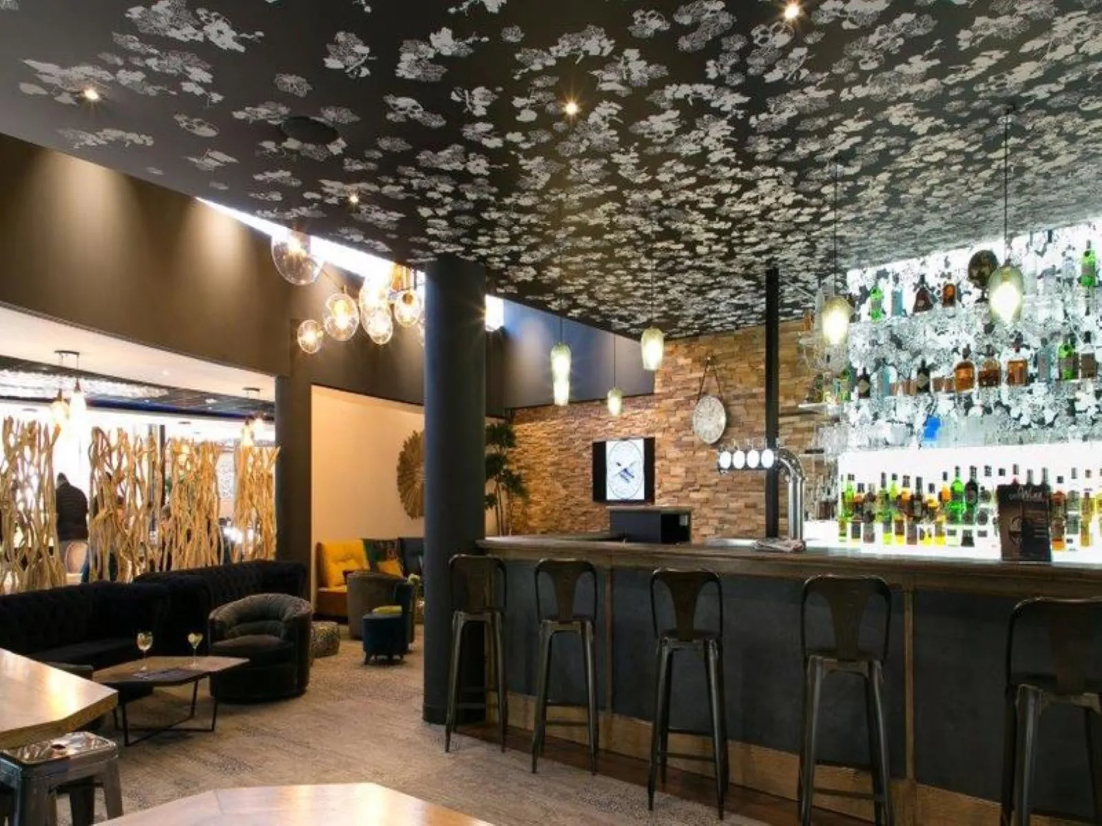 Elegant bar at Mercure Bordeaux Airport Hotel featuring a decorative floral patterned ceiling, black counter, bar stools, modern furniture, pendant lighting, and exposed brick walls