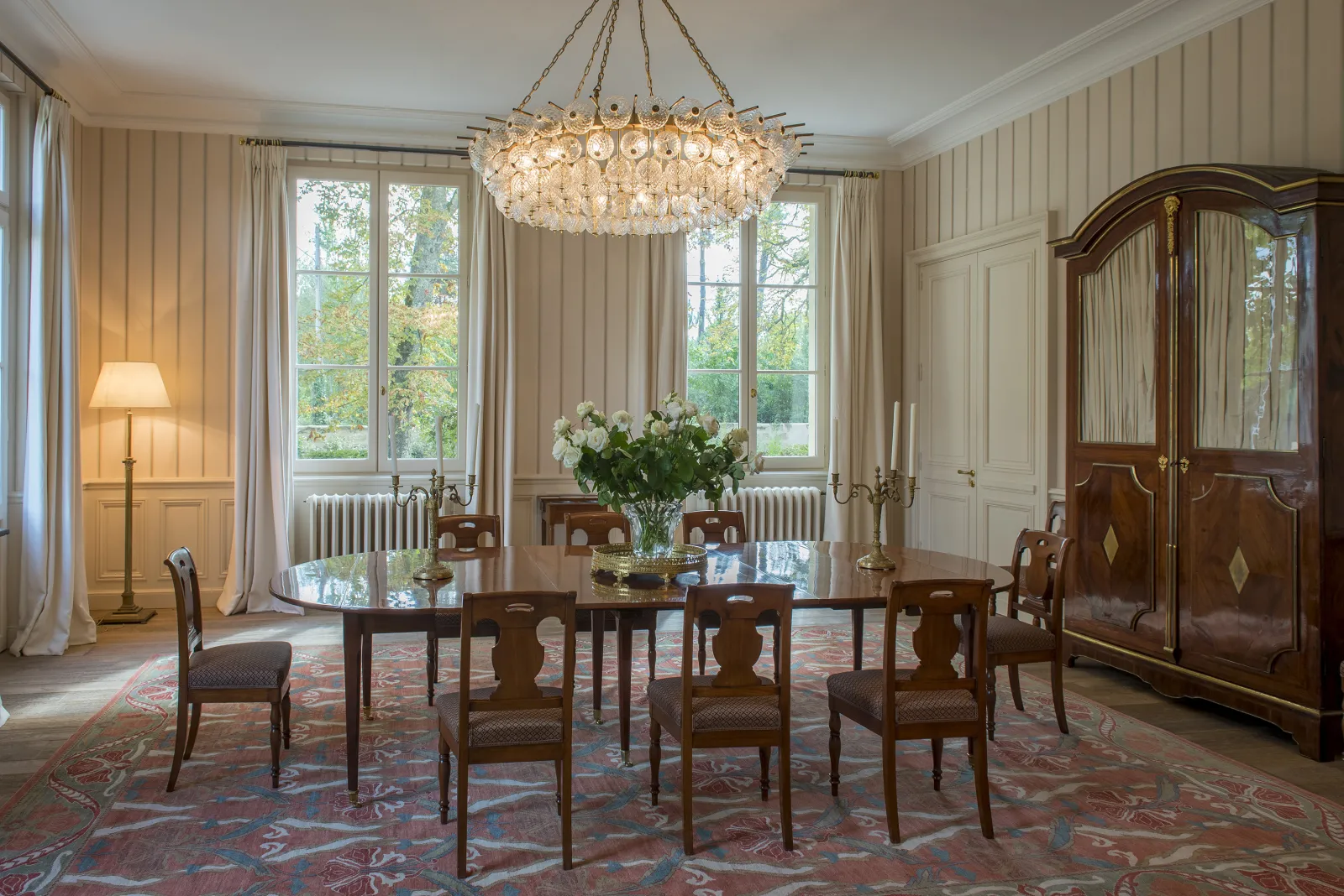 Elegant dining room featuring a dark wood table with matching chairs, crystal chandelier, tall windows with beige curtains, wooden display cabinet, and white floral centerpiece