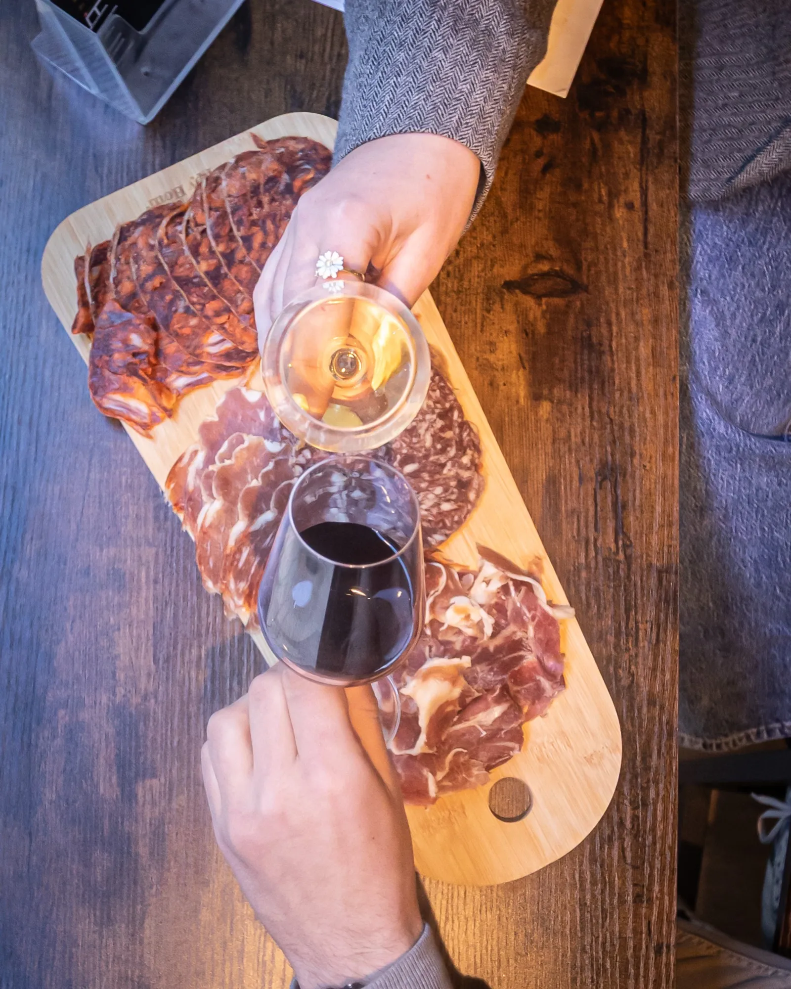 A person wearing a diamond ring holds two wine glasses (white and red wine) above a wooden board with cured meats and aged cheeses.