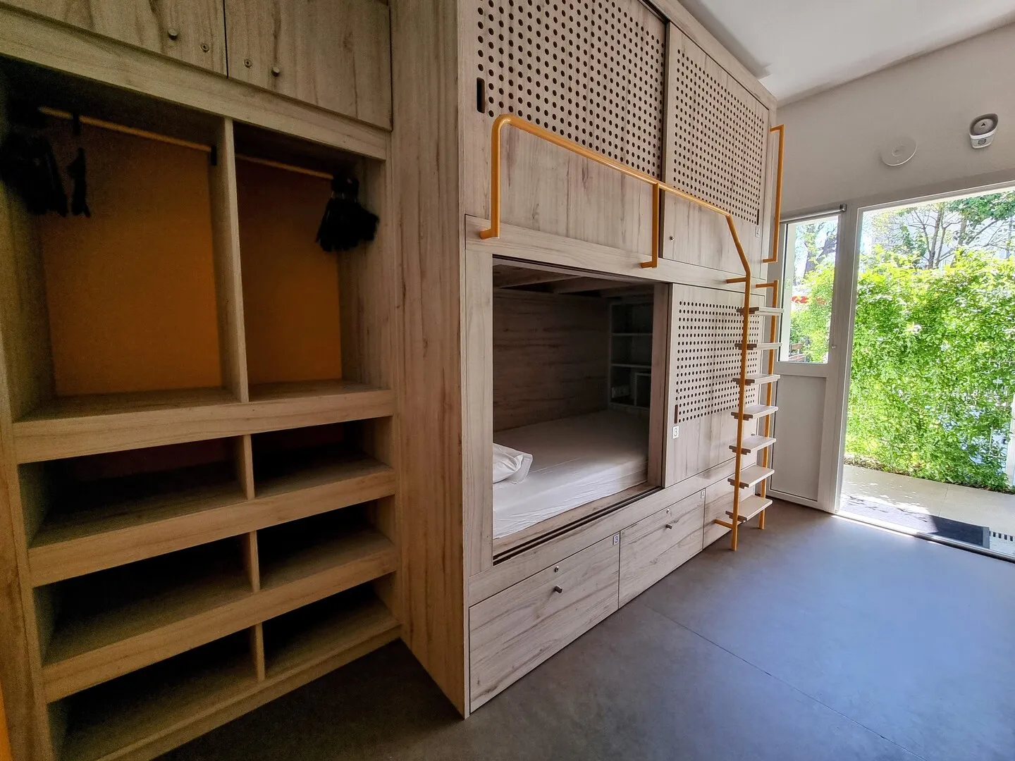 Modern hostel room featuring a light wood bunk bed with storage compartments, open shelving, wooden shutters, and glass door overlooking a green garden in Bordeaux.