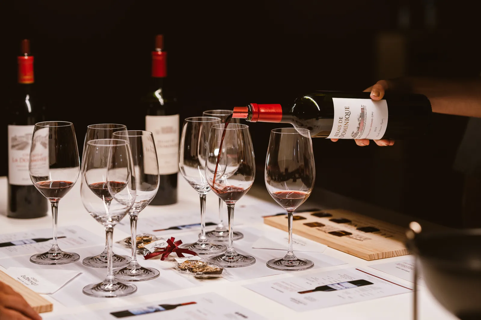 Wine tasting in Bordeaux: red wine glasses filled with Bordeaux wine, bottles with visible labels, lit red candles, tasting notes on white table, and a hand pouring wine.