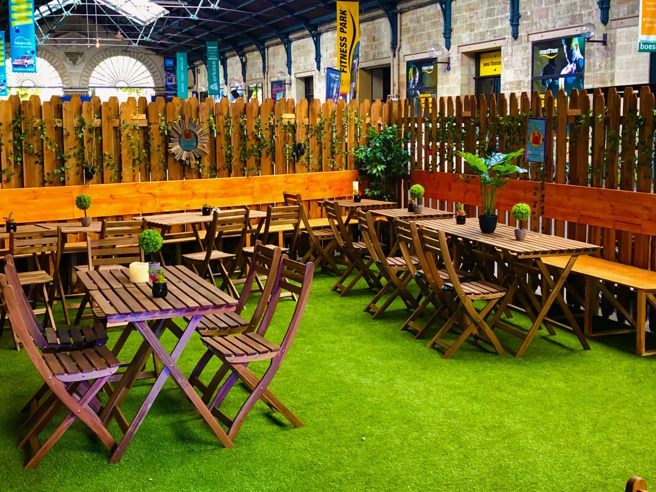 Indoor relaxation area at Théâtre des Chartrons in Bordeaux featuring artificial grass, colorful wooden deck chairs, high tables, decorative fencing, and signage, under a covered structure with historic architecture.