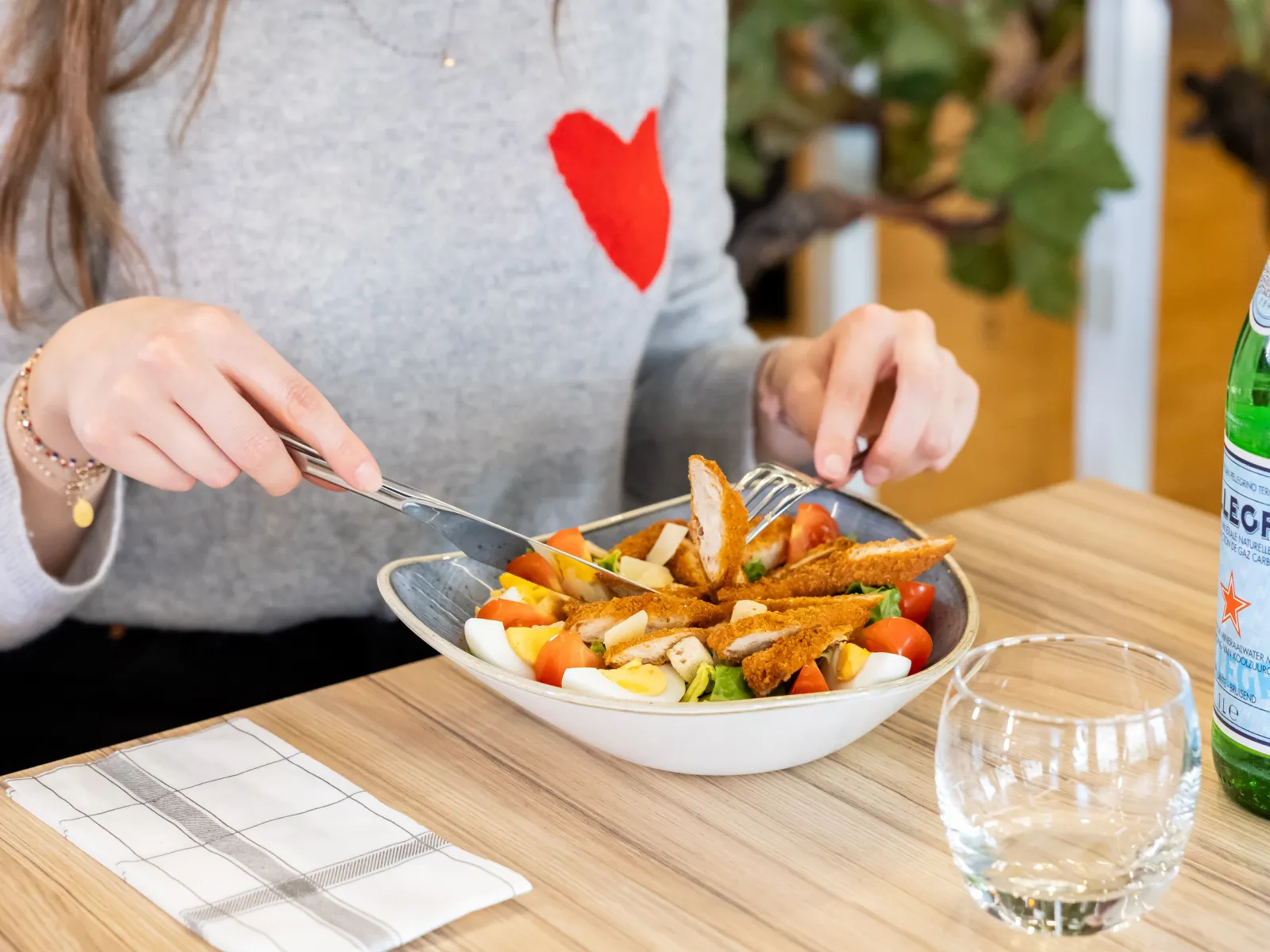 A woman wearing a red heart on her chest enjoys a plate of fries and vegetables at a bistro, with a glass of water and a beer nearby.