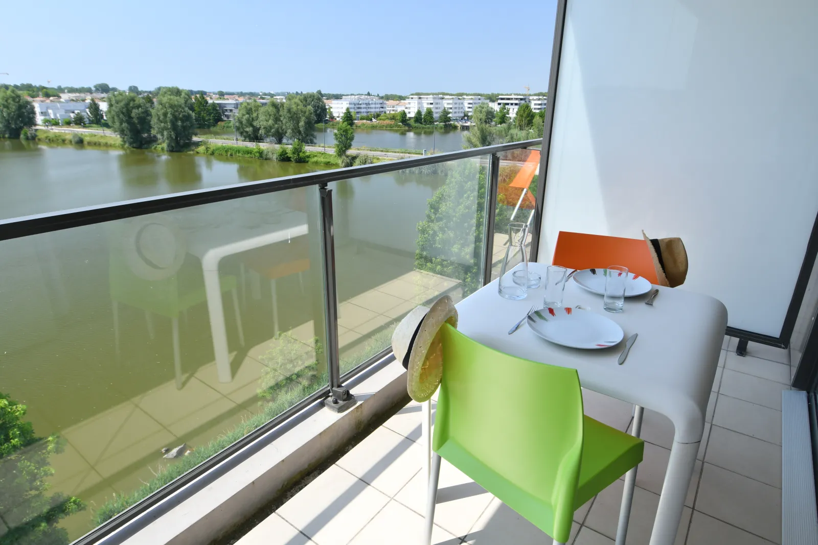 Balcony overlooking the Garonne river in Bordeaux, set dining table with green and orange chairs, water glass and plates, with residential buildings visible across the river