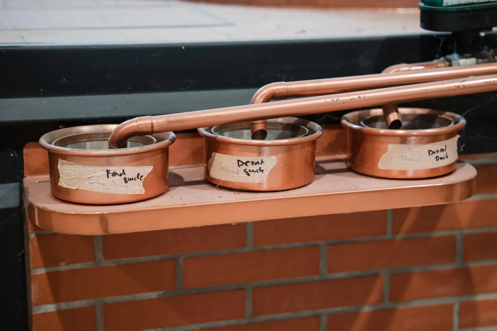 Three copper stills lined up on a platform, labeled and connected by copper pipes, at Citadelle Gin Distillery in Bordeaux
