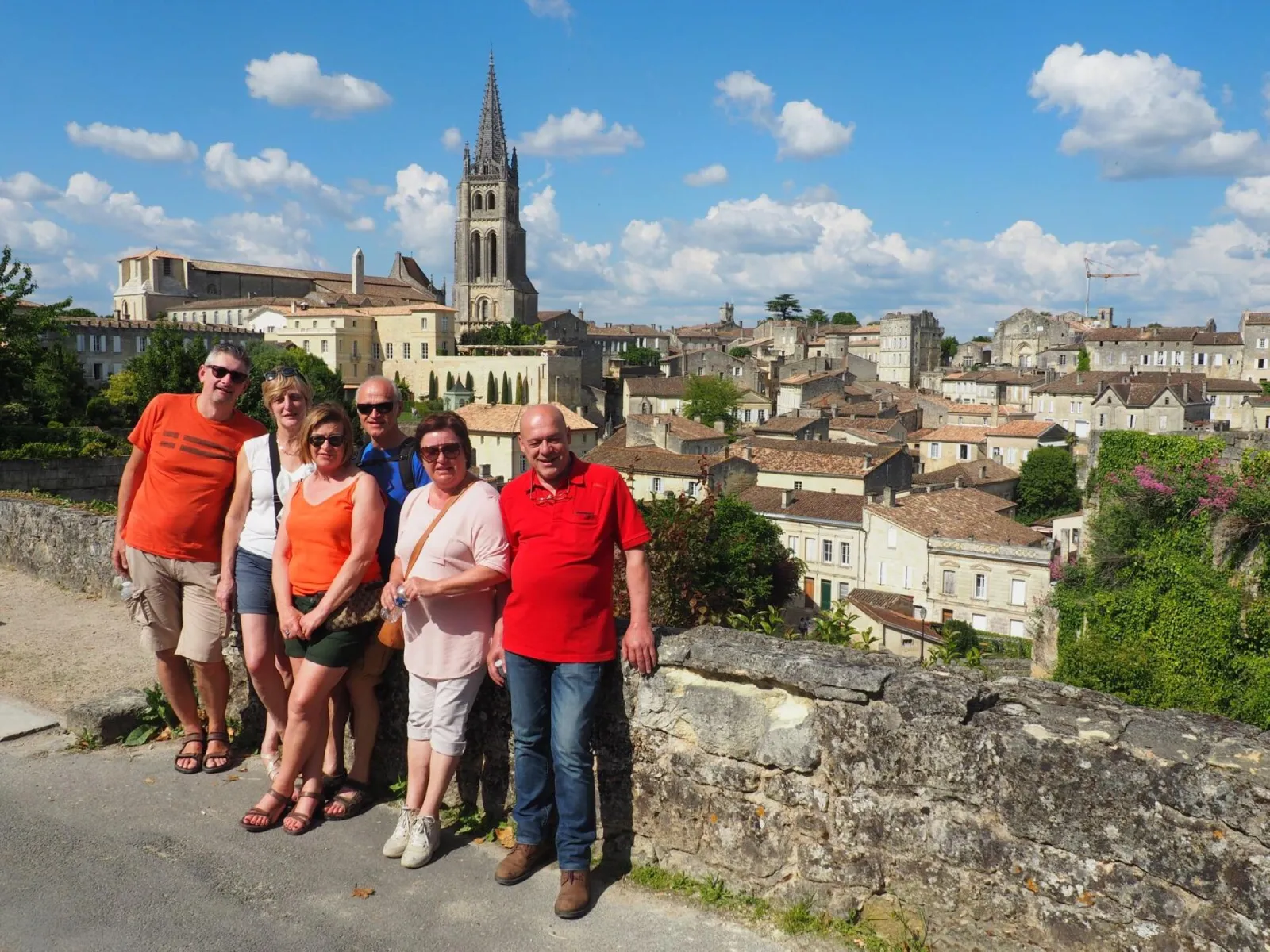 A group of six tourists posing in front of a panoramic view of a medieval town with a Gothic church dominating the horizon, stone houses and cloudy blue sky.