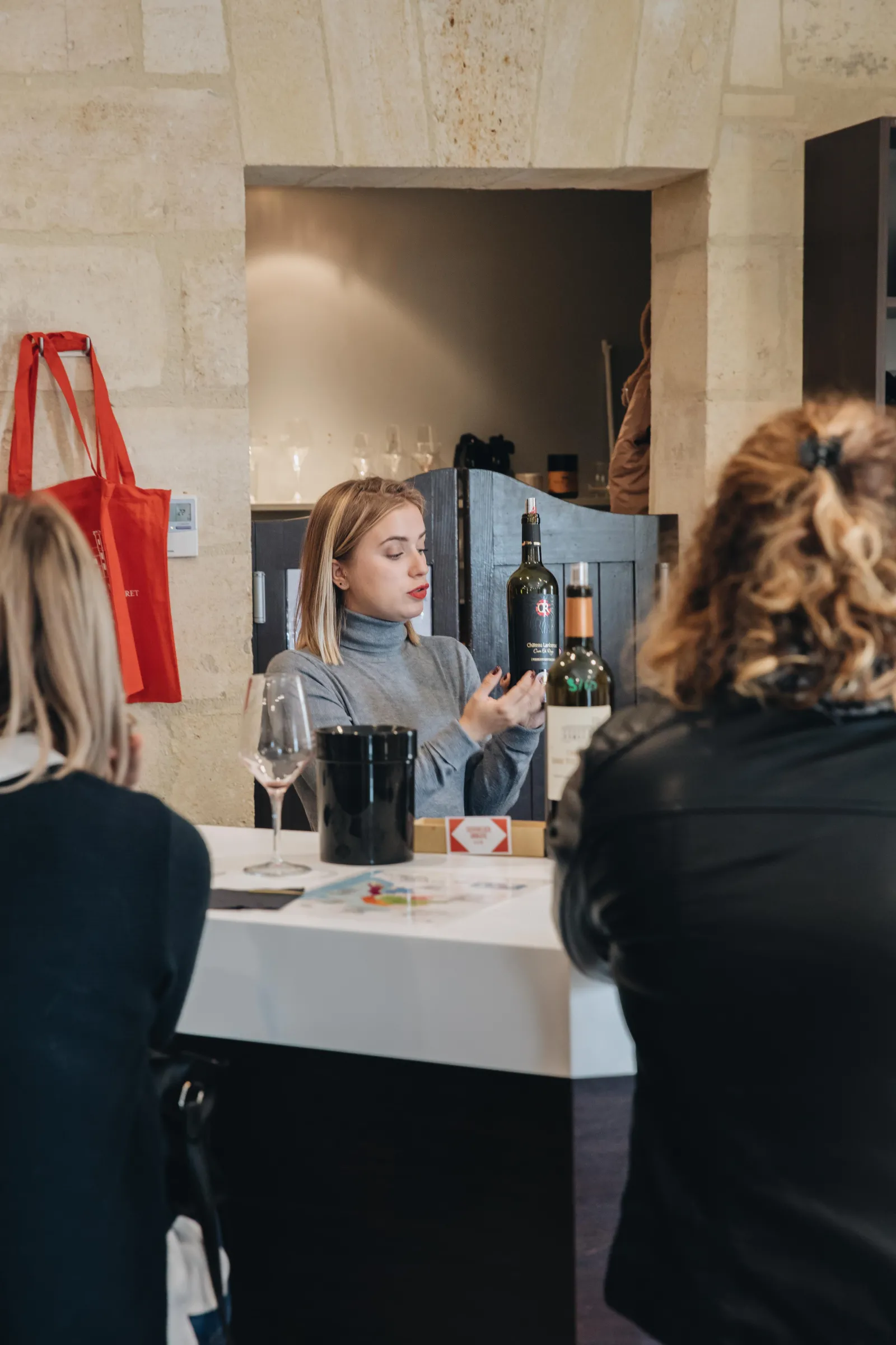 Wine tasting at Château Ambe Tour Pourret in Bordeaux: a woman presents wine bottles to two visitors around a white table.