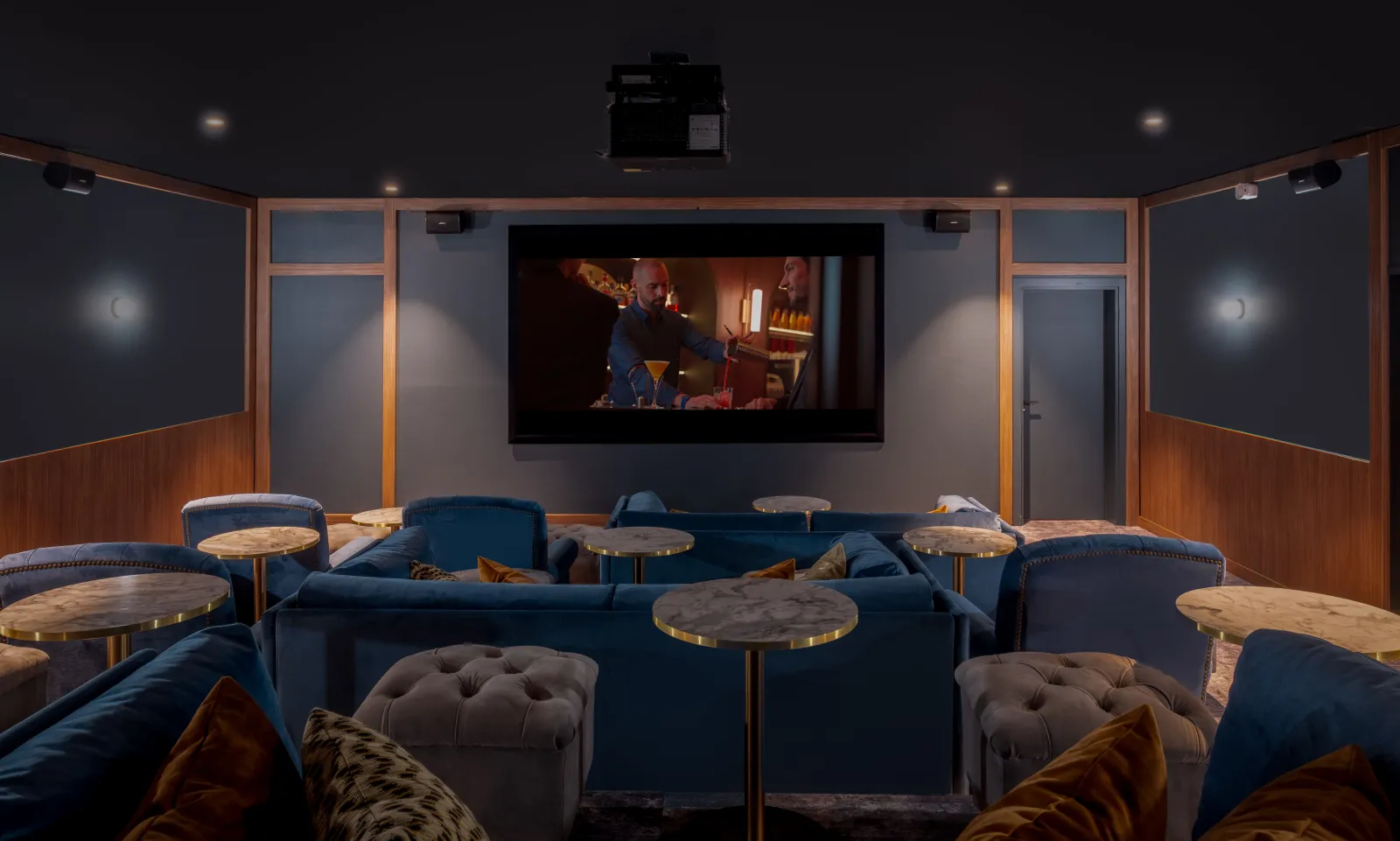 Luxury private cinema room featuring blue velvet seating, marble and brass side tables, wall-mounted screen displaying a film, and elegant wood paneling.