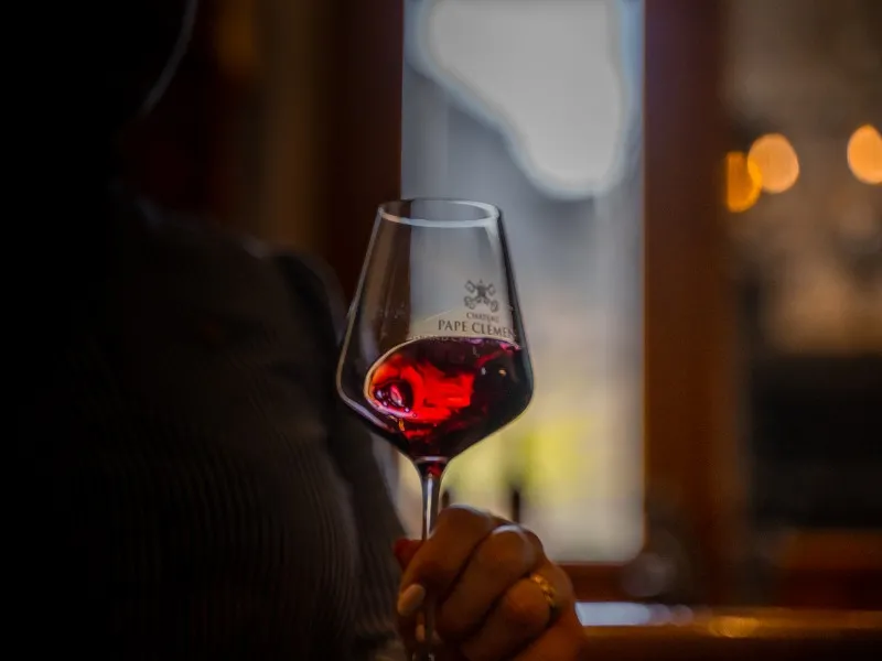 Glass of red wine from Château Pape Clément held in hand against a window overlooking Bordeaux, with warm evening lighting.