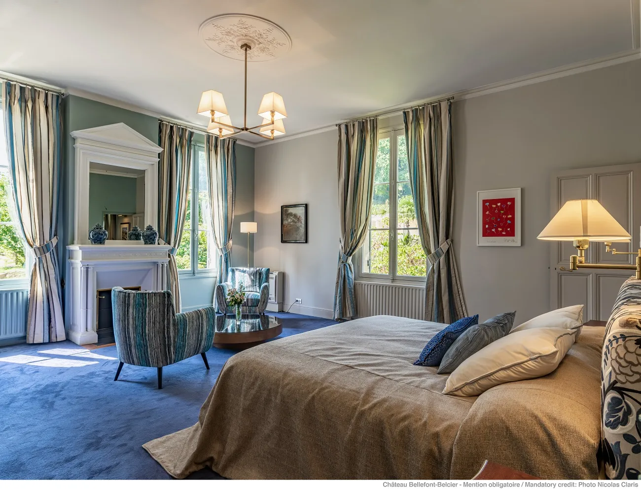 Luxury bedroom suite at Château Bellefont-Belcier in Bordeaux, featuring a king-size bed, marble fireplace, striped blue armchairs, draped curtains, bronze chandelier, and windows overlooking the estate.