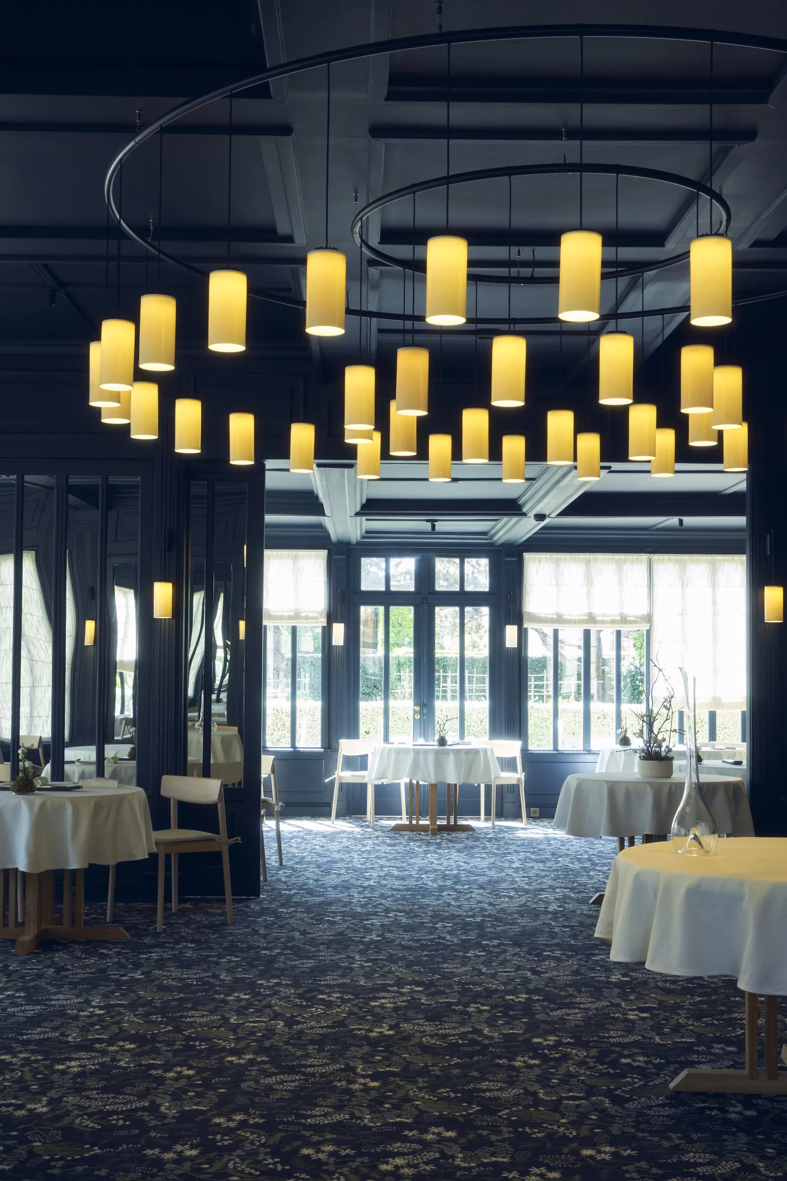 Elegant restaurant dining room featuring a modern chandelier with yellow cylindrical lights, round white tables, large windows, and contemporary décor in Bordeaux
