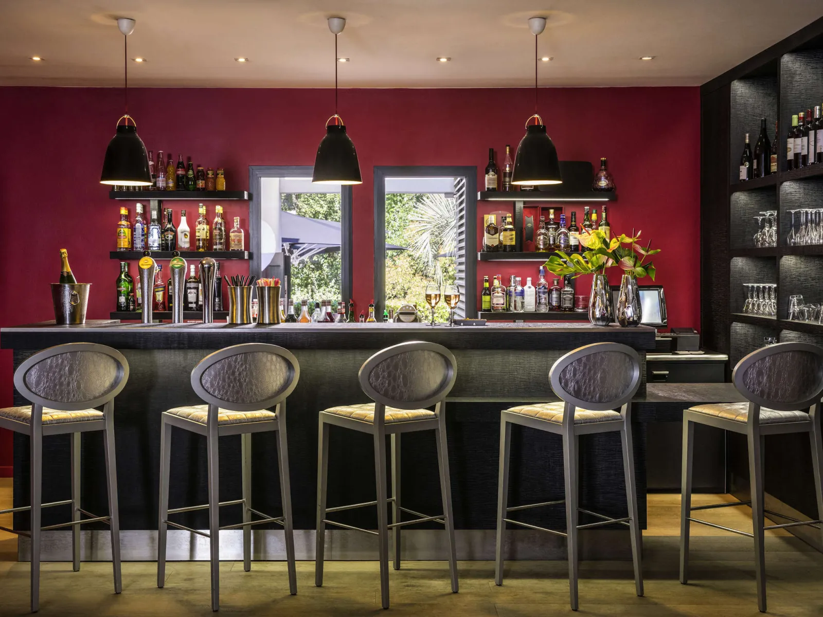 Elegant bar with burgundy red walls, black counter, five gray bar stools, three black pendant lights, shelves of alcohol bottles, and views of nature through glass doors