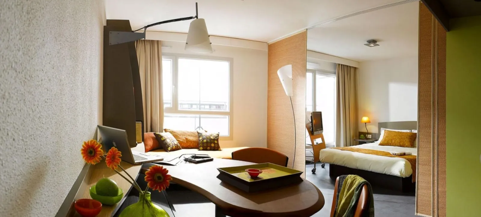 Spacious hotel suite at Adagio Bordeaux featuring a work area, orange sofa with cushions, white desk, fresh flowers, and bedroom with double bed and large windows in the background.