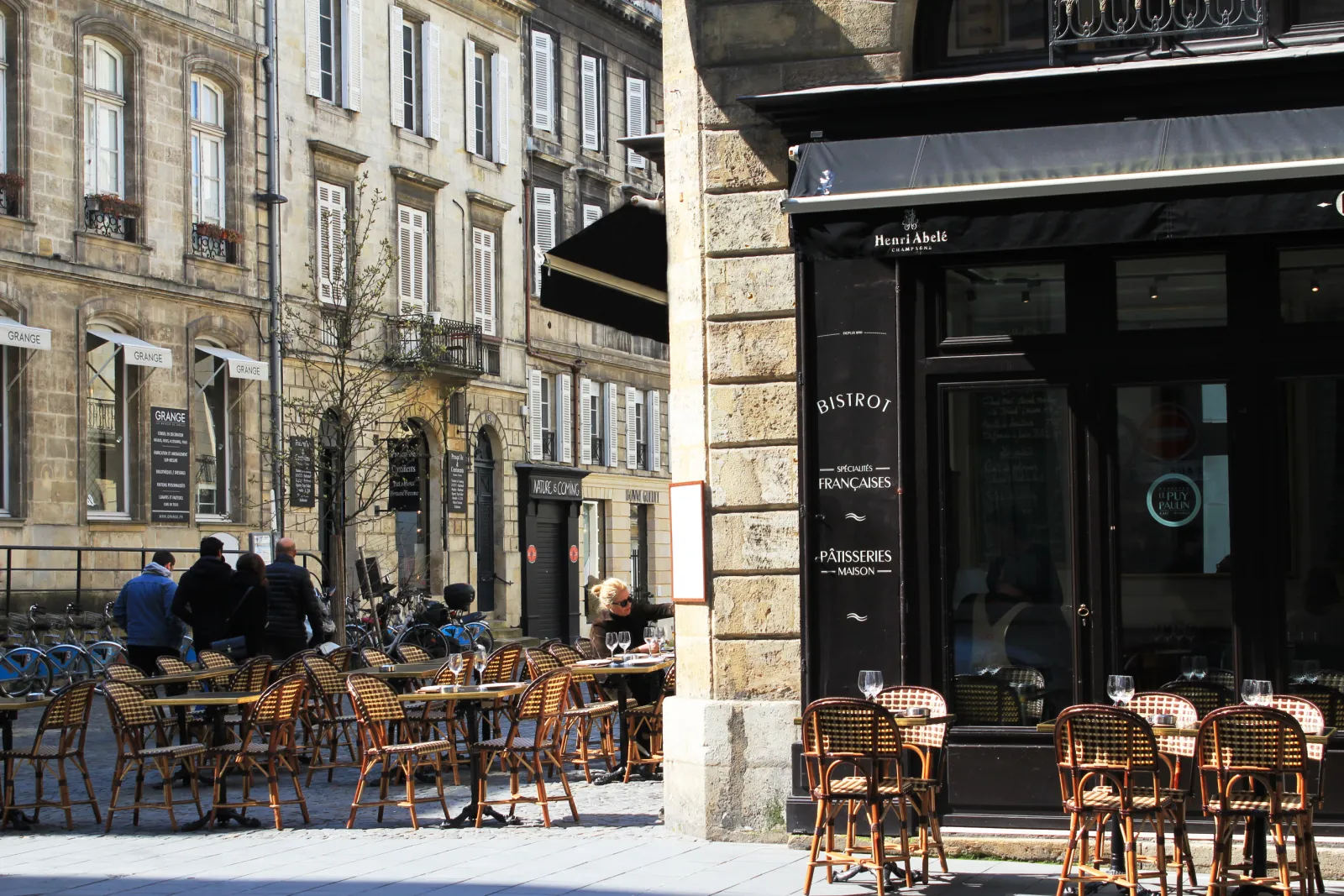 Outdoor terrace of a French bistro and Henri Abélé bakery on a paved street in Bordeaux, featuring rattan chairs and traditional stone facades in the Puy-Paulin district