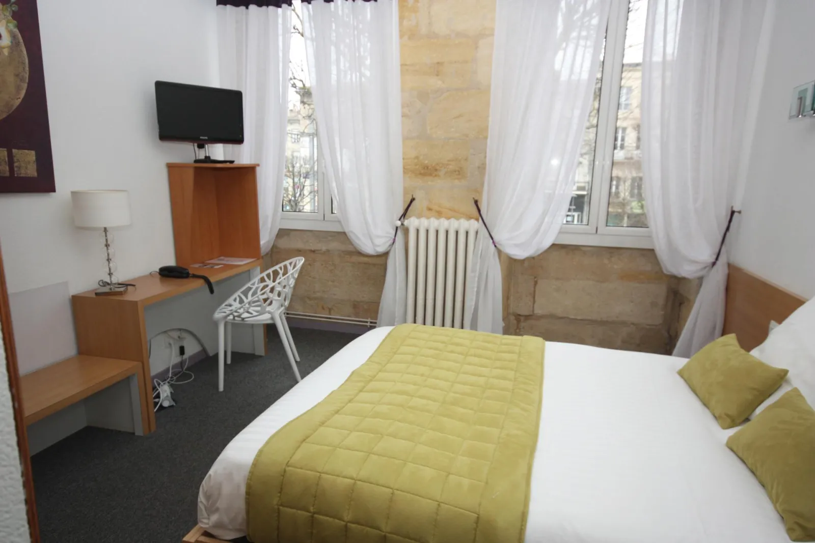 Hotel room in Bordeaux featuring a white bed with yellow bedspread, wooden desk, white chair, television, radiator and windows with white curtains overlooking the city