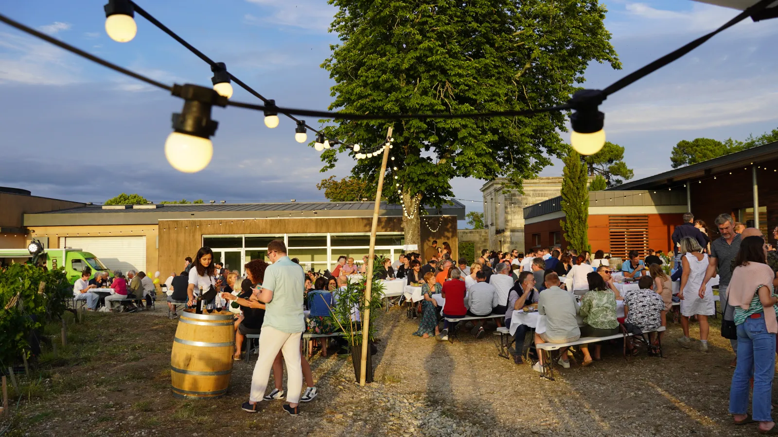 Outdoor event at Château Luchey-Halde in Bordeaux featuring guests seated at tables, string lights, a wine barrel, and modern buildings in the background.