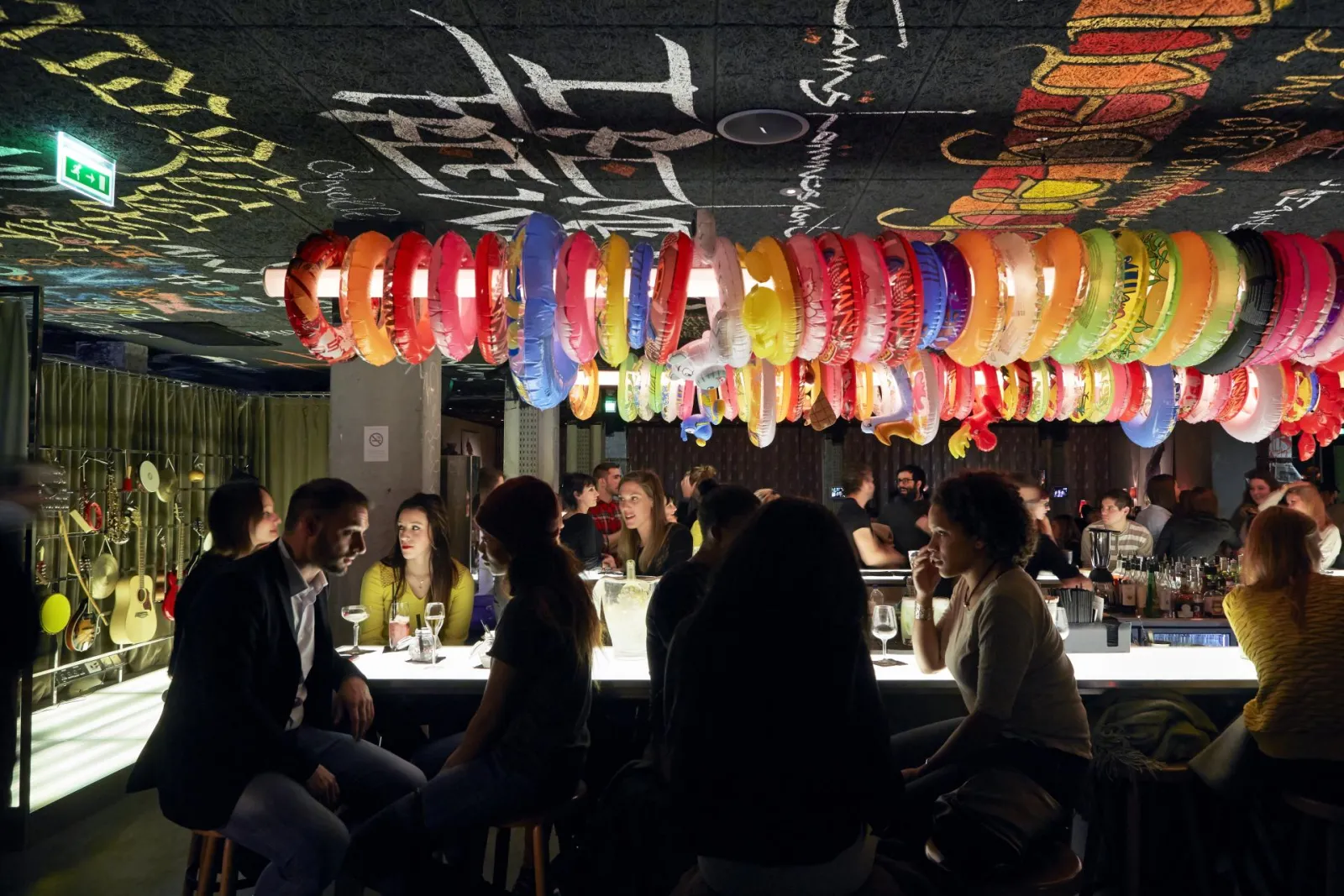 Interior of Mama Shelter bar in Bordeaux with customers seated at an illuminated counter, beneath a ceiling decorated with colorful bracelets and urban art.