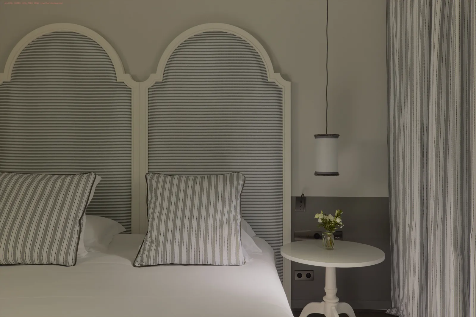 Elegant bedroom featuring two arched gray slatted headboards, striped pillows, white round nightstand with flower vase, vertical striped curtains, and modern pendant light.