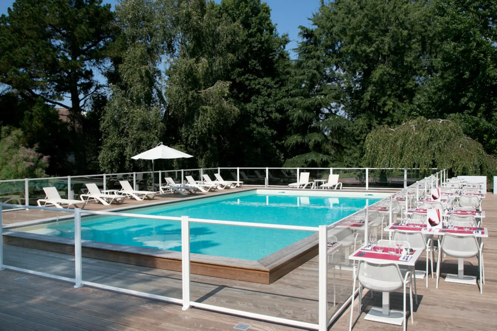 Outdoor swimming pool at Mercure Bordeaux-Lac hotel featuring lounge chairs, white umbrella, wooden deck, dining tables with red and white chairs, surrounded by lush greenery.