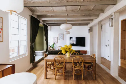 Bright loft interior with exposed wooden beams, wooden dining table with rattan chairs, large windows, open kitchen, and minimalist décor in Bordeaux.