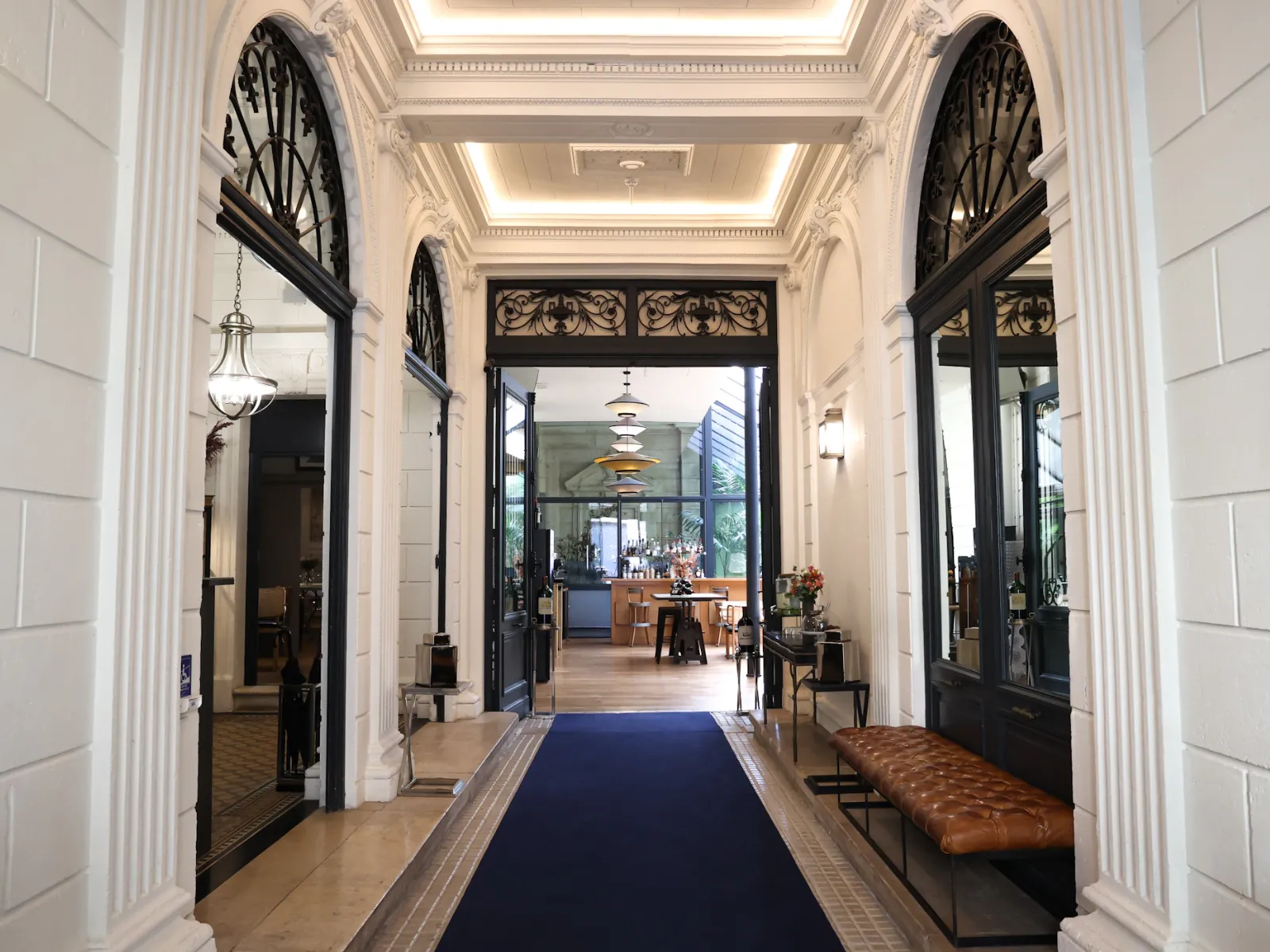 Elegant entrance hallway of a Bordeaux boutique hotel featuring white columns, ornate black doors, coffered ceiling with lighting, and blue carpet leading to the reception area