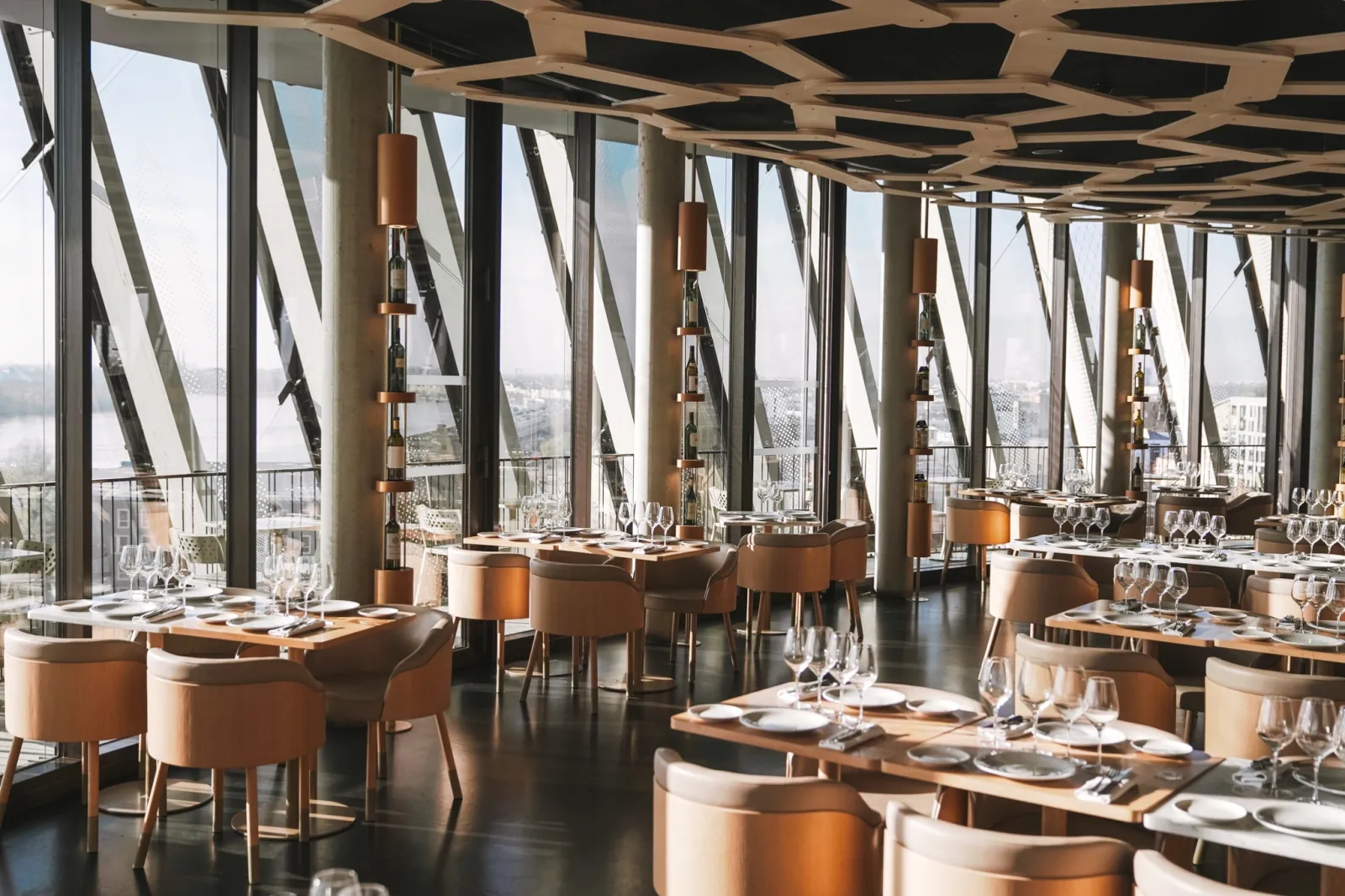 The 7 panoramic restaurant at La Cité du Vin in Bordeaux featuring floor-to-ceiling windows overlooking the city and Garonne river, set dining tables with cognac leather chairs, and modern geometric ceiling design