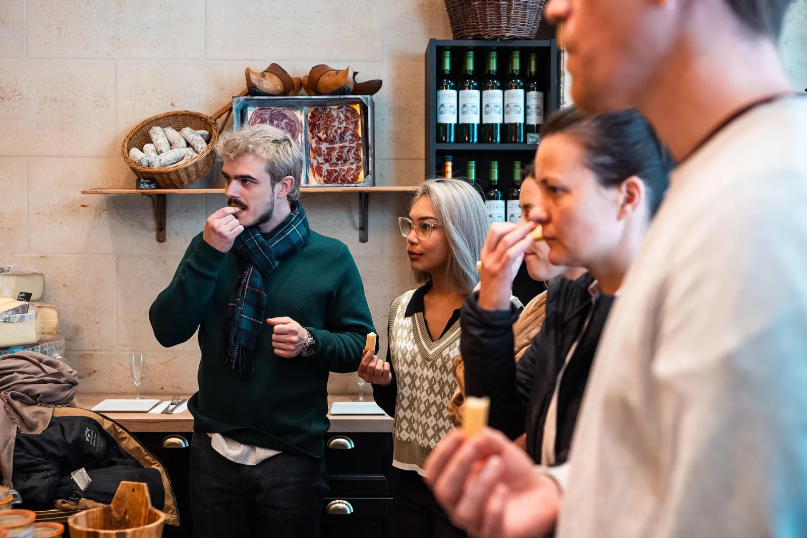 Food tour guide presenting cured meats and wines to visitors in Bordeaux, standing in front of shelves displaying Iberian ham, bread, and bottles of red wine.