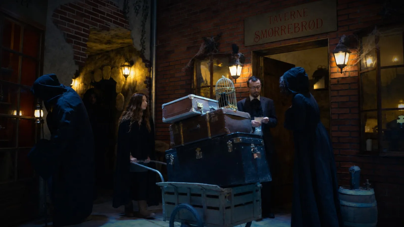 Nighttime scene in a Bordeaux alley with figures in dark clothing gathered around trunks and a birdcage in front of an illuminated tavern storefront