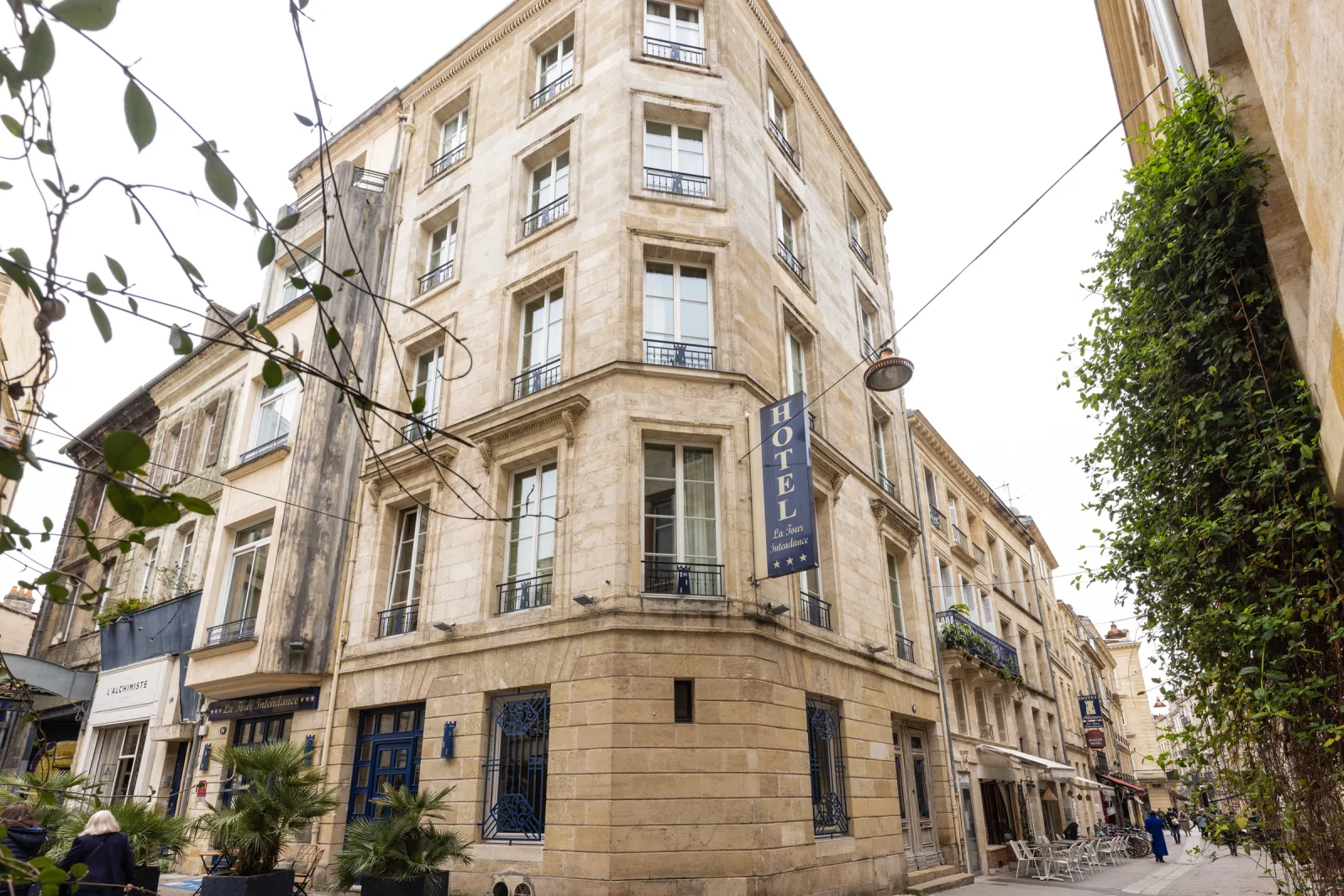 Facade of the Hôtel de la Tour Intendance in Bordeaux, a Haussmann-style building with ashlar stone and wrought iron balconies, blue hotel sign visible on the street corner, surrounded by vegetation and shops.