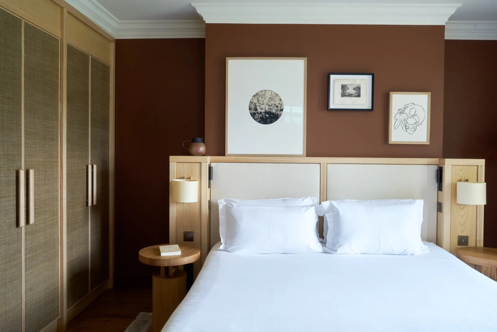 Elegant bedroom with terracotta walls, king-size bed with white linens, light wood headboard with integrated niches and lamps, built-in wardrobe on the left, and three framed artworks above the bed.