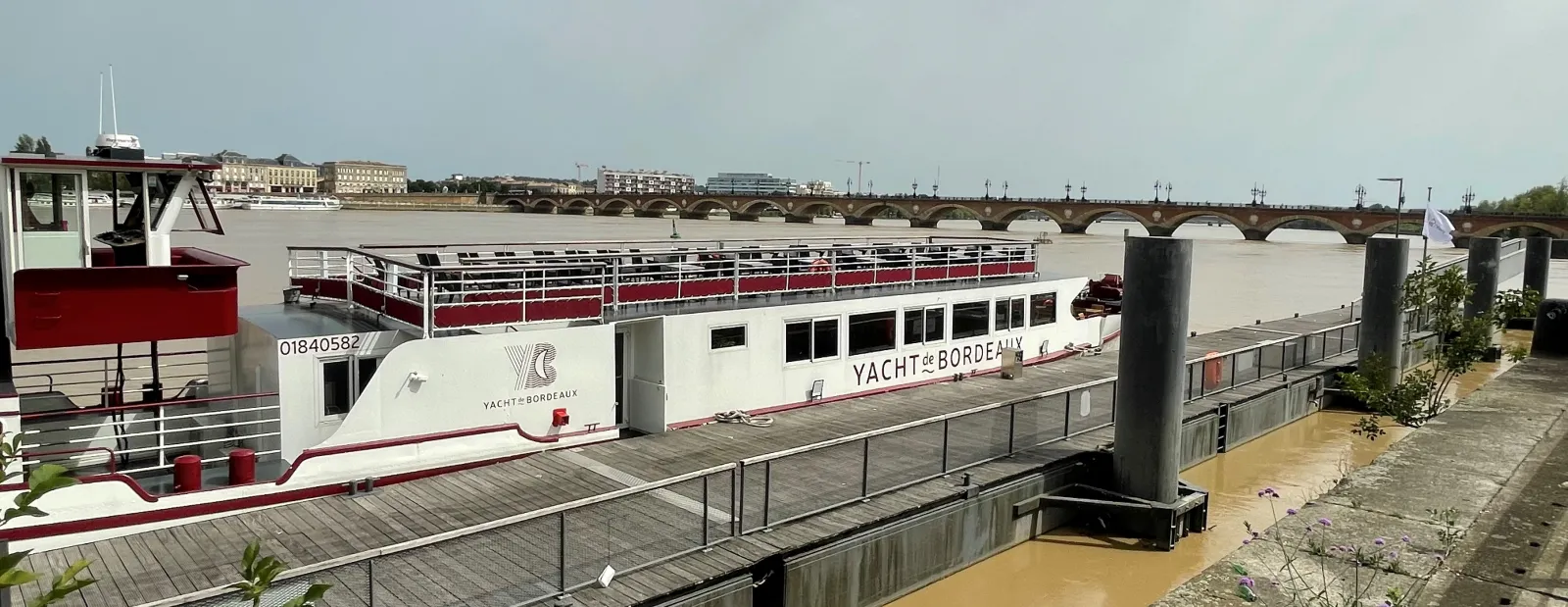 The Maddalena, a white and red river cruise yacht, moored at a Bordeaux quay with the Garonne river and Pont de Pierre bridge visible in the background.