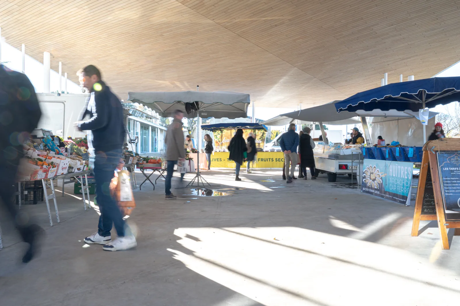 Covered market in Bouliac, Bordeaux featuring produce stalls, shoppers browsing under the Vettiner hall, natural light streaming through the structure.