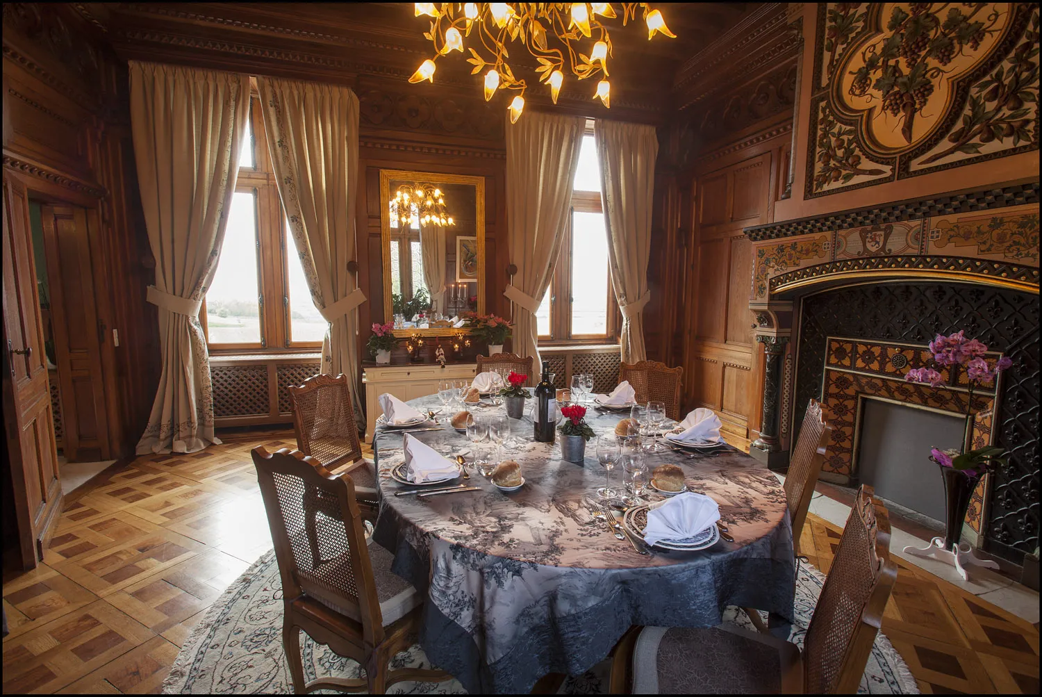 Elegant dining room at Château de La Rivière featuring a round table, golden chandelier, ornate fireplace and windows overlooking the Bordeaux countryside