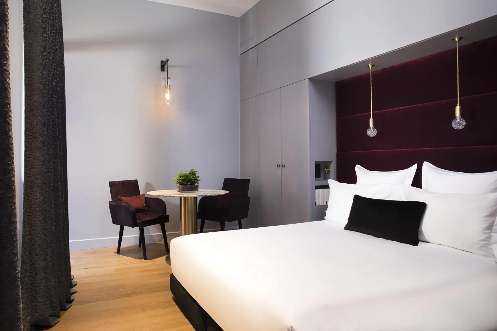 Modern hotel room at Cardinal Bordeaux featuring a white bed with burgundy headboard, round work desk with chairs, gold pendant lights, and wooden flooring.
